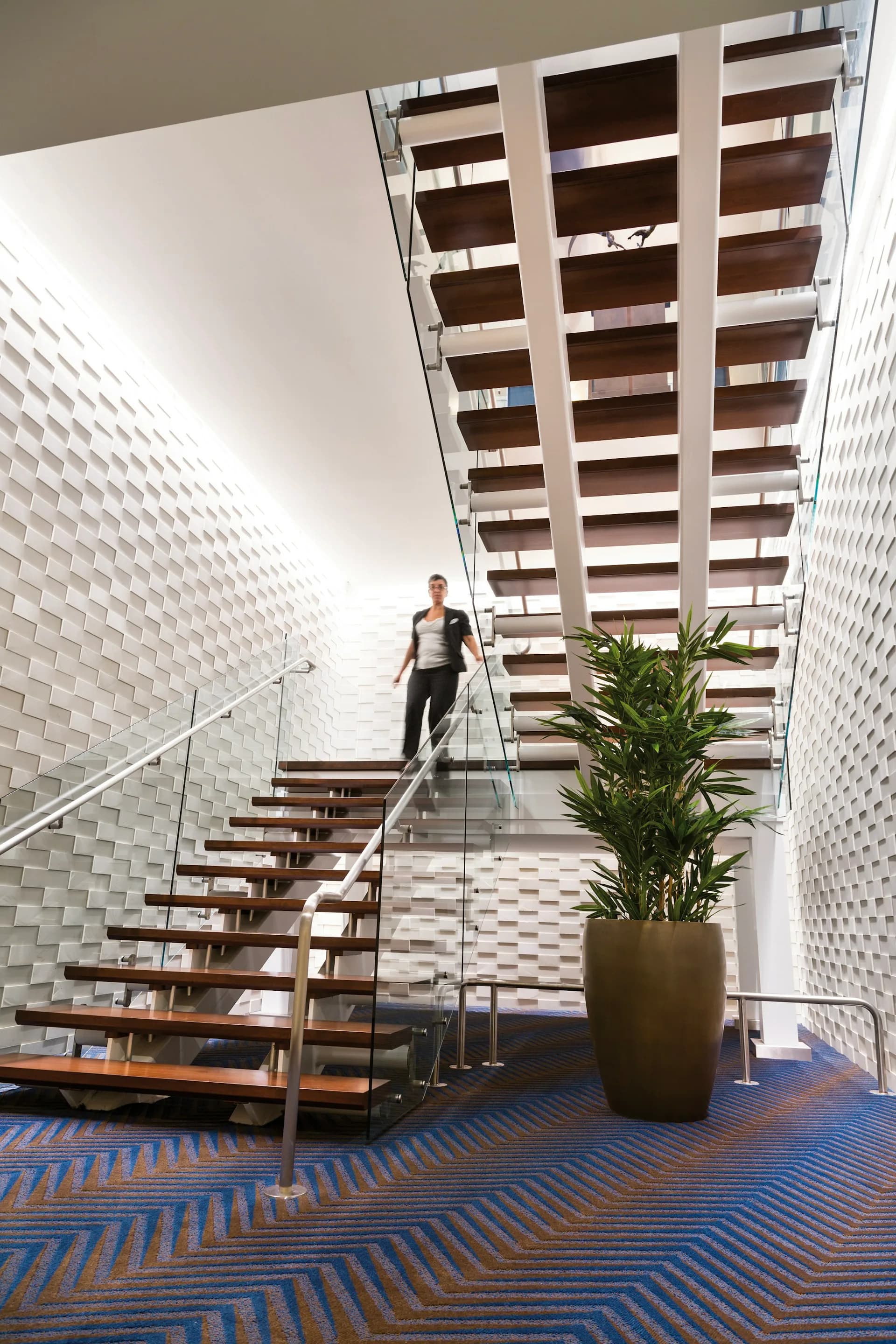 Modern atrium staircase with floating wood steps, glass railings, textured white brick walls, potted plant, and patterned blue flooring designed by Cooper Carry.