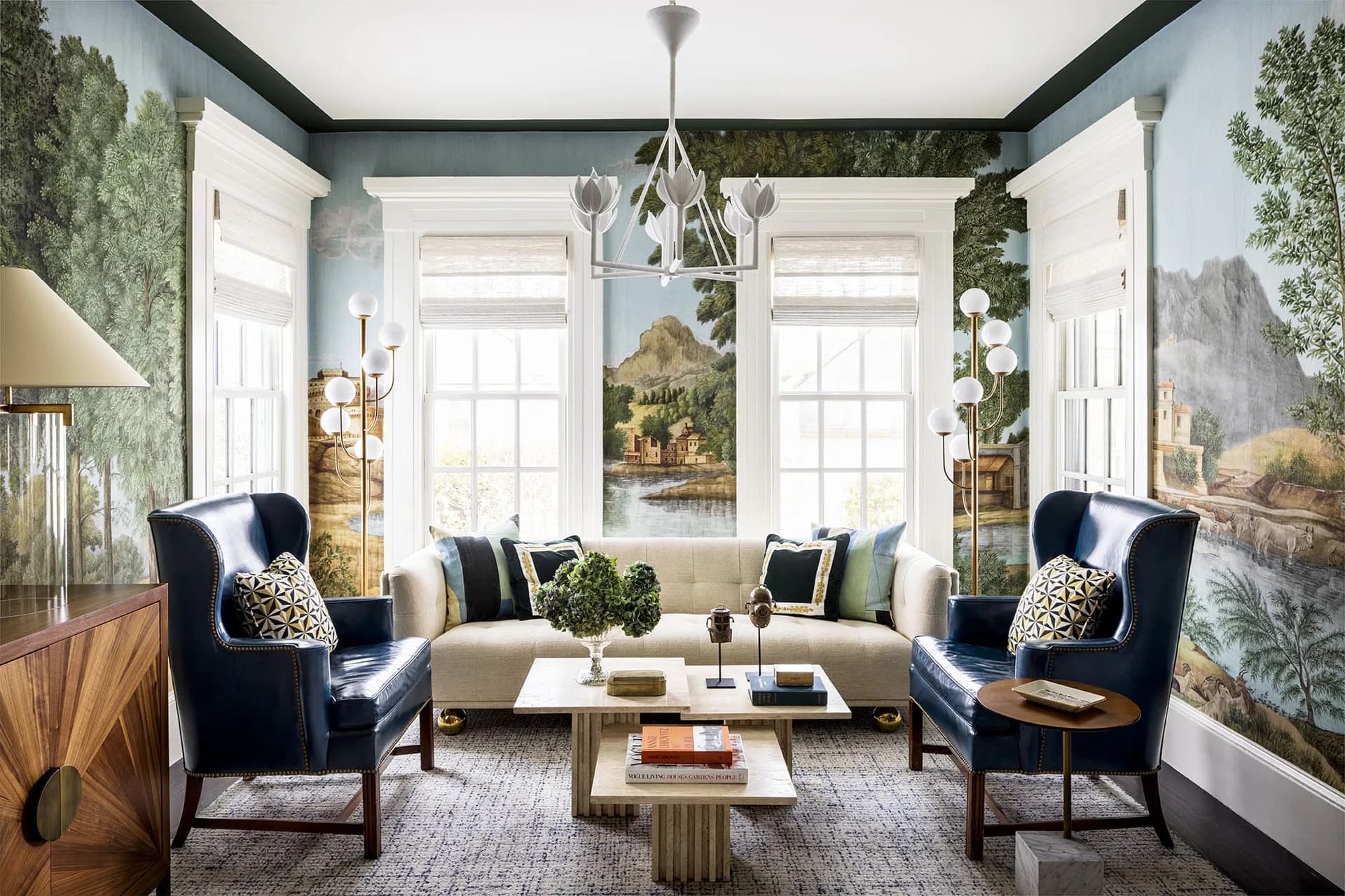 Formal living room with navy wingback chairs, cream sofa, landscape wallpaper, large windows, and brass chandelier by Corey Damen Jenkins & Associates.