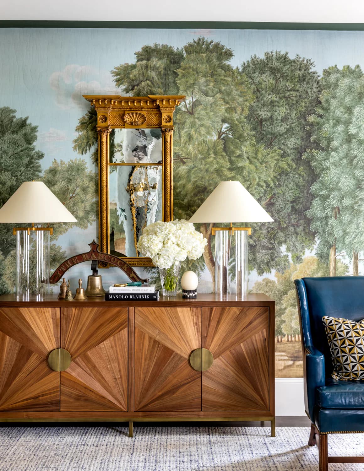 Sophisticated living room featuring a wood console with sunburst inlay doors, ornate gold mirror, white lamps, and lush forest wallpaper backdrop by Corey Damen Jenkins & Associates.