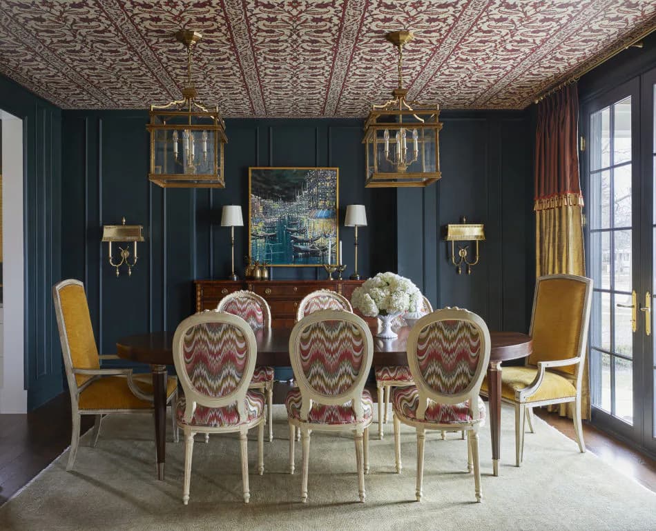 Formal dining room with dark teal walls, ornate ceiling, brass pendant lights, and chevron-patterned upholstered chairs around a dark wood table by Corey Damen Jenkins & Associates.