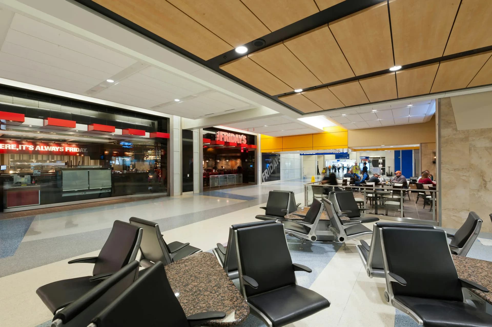Modern airport terminal lounge with black seating, wood ceiling panels, neutral flooring, and commercial storefronts by Corgan.