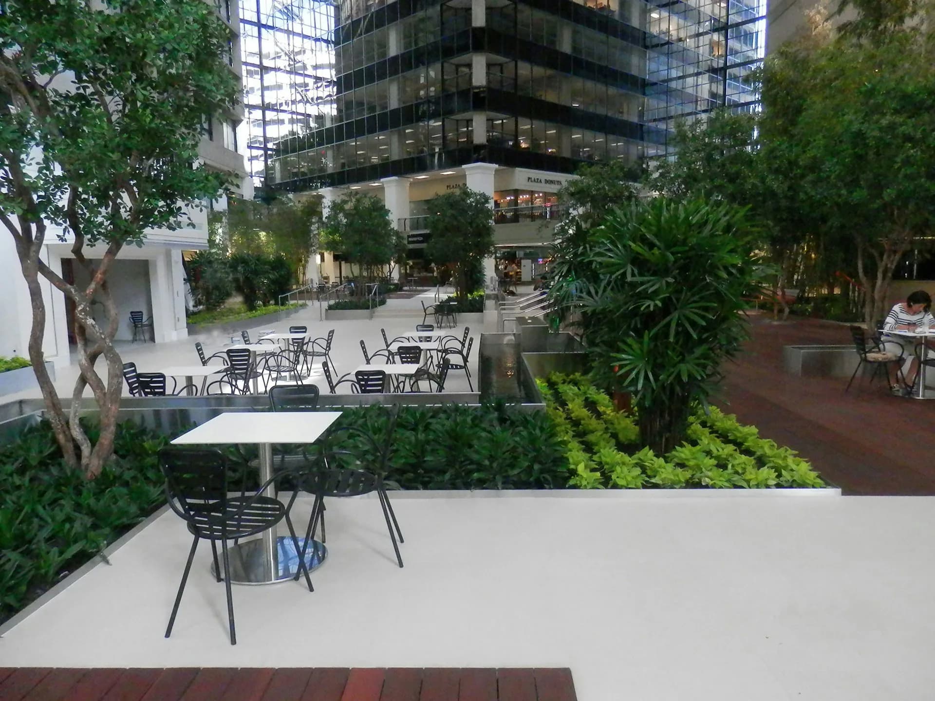 Modern outdoor plaza with white flooring, contemporary seating areas, lush green landscaping, and tall buildings in background, designed by Corgan.