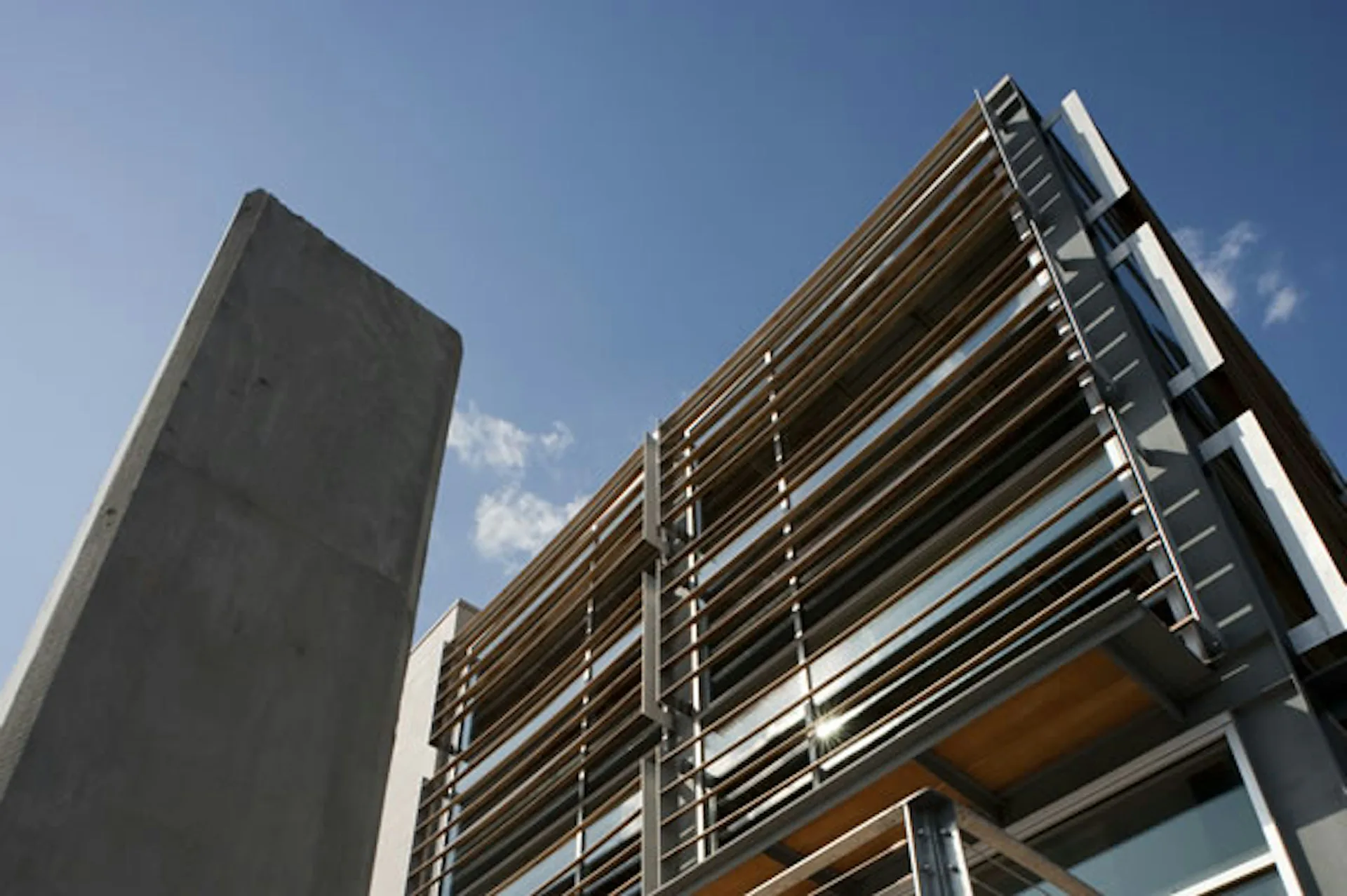 Contemporary mixed-use building exterior featuring a dark concrete tower alongside a multi-story structure with horizontal metal louvers and glass panels against a blue sky, by CREATE Architecture Planning & Design.