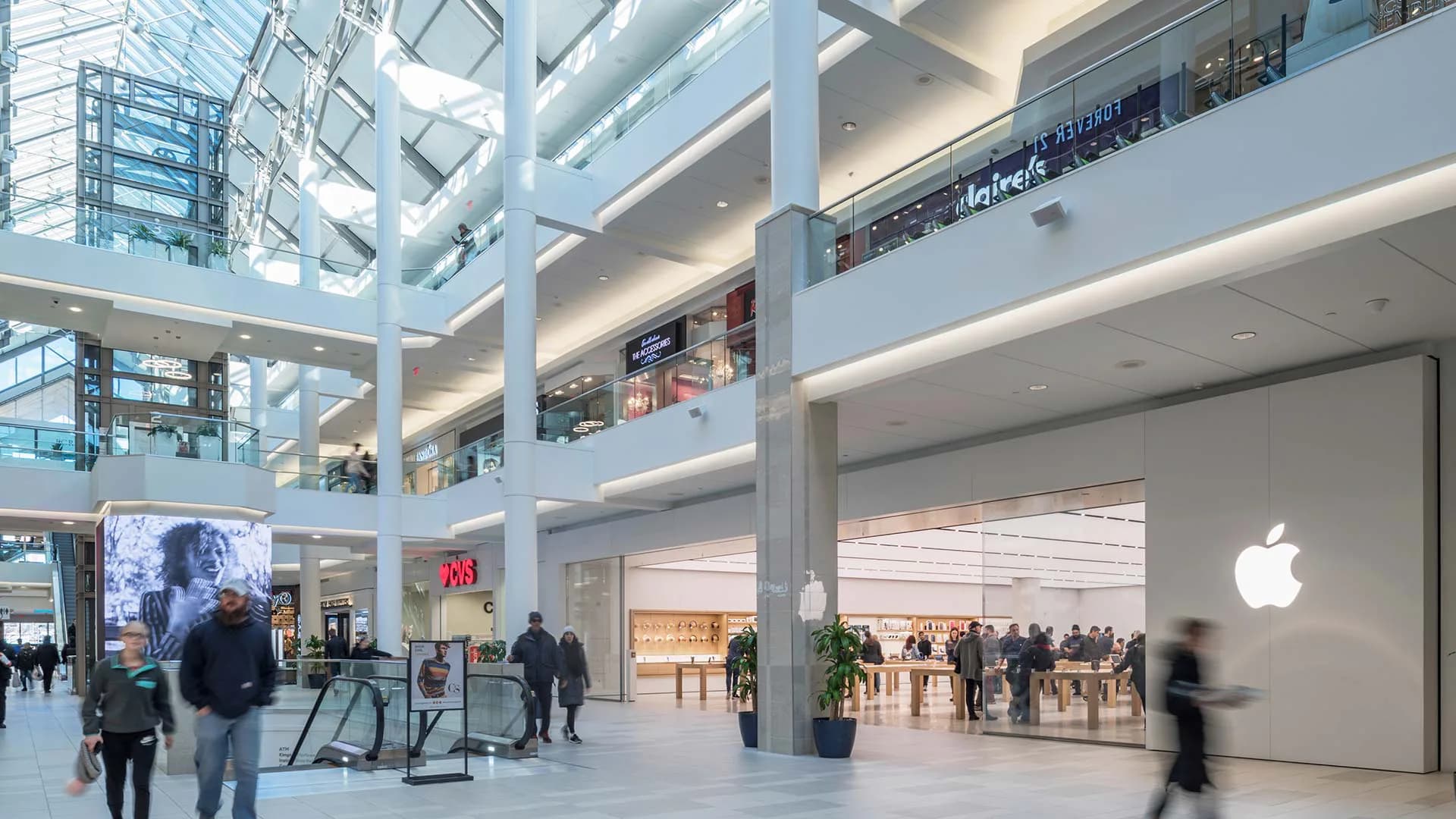 Multi-level modern shopping mall interior with bright skylight, glass railings, and Apple retail storefront by CREATE Architecture Planning & Design.