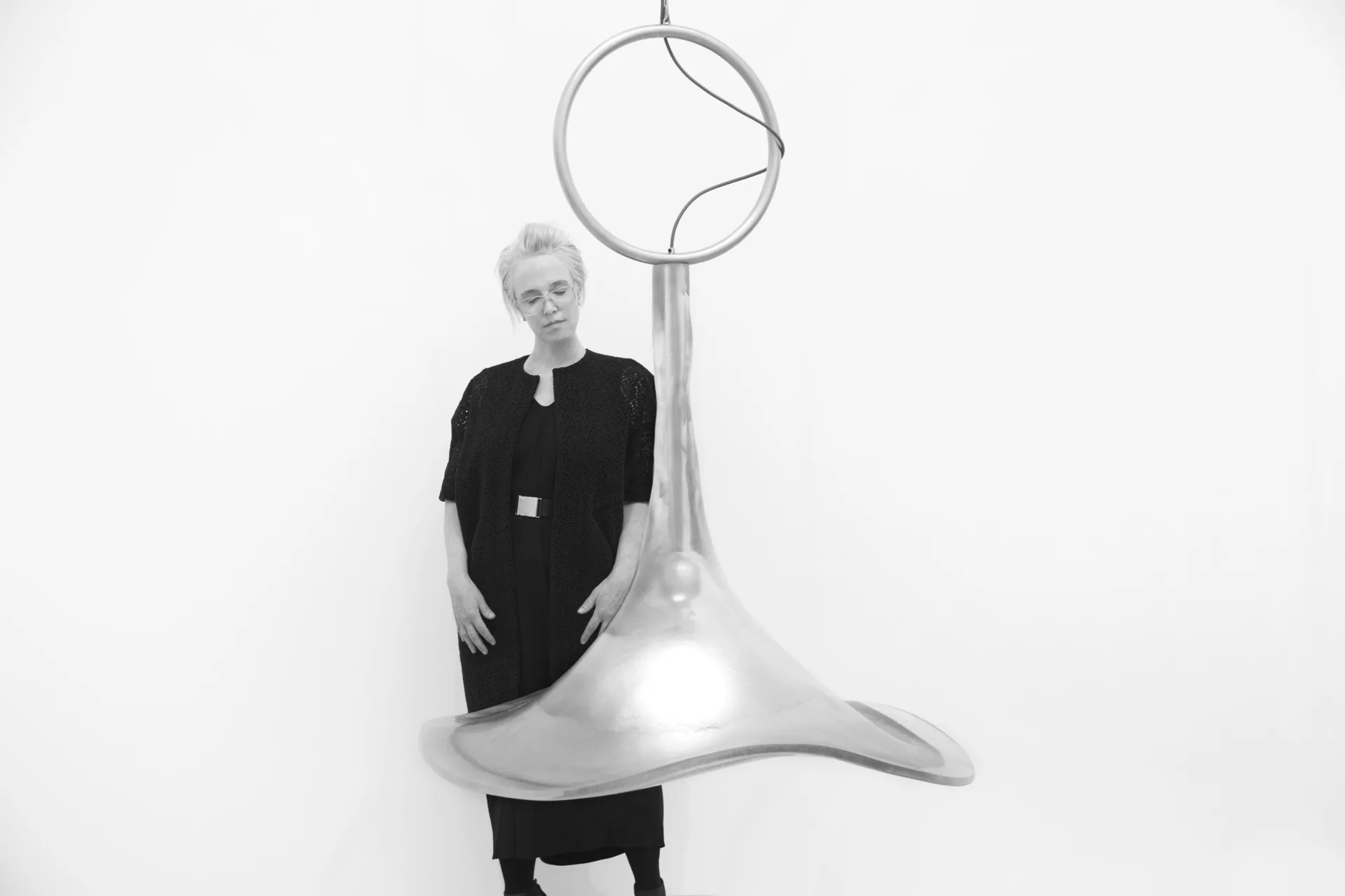 A person in black clothing poses with a sculptural installation featuring a large silver funnel-like form with a geometric circle mounted above, creating a striking three-dimensional art piece by Cristina Grajales Gallery.