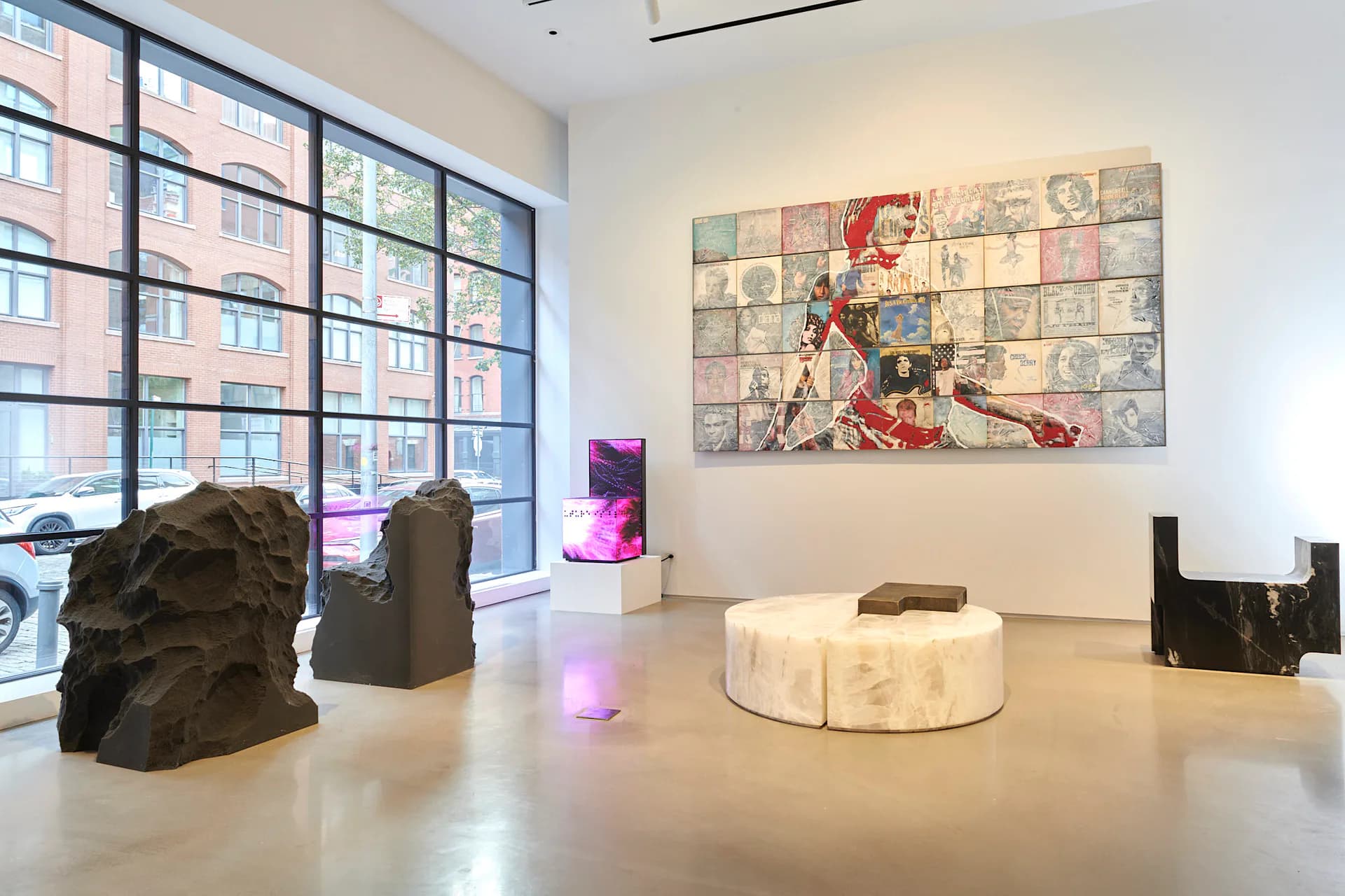 Contemporary gallery interior with polished concrete floor, sculptural dark installations, circular white plinth, colorful comic-style wall artwork, and expansive grid windows by Cristina Grajales Gallery.