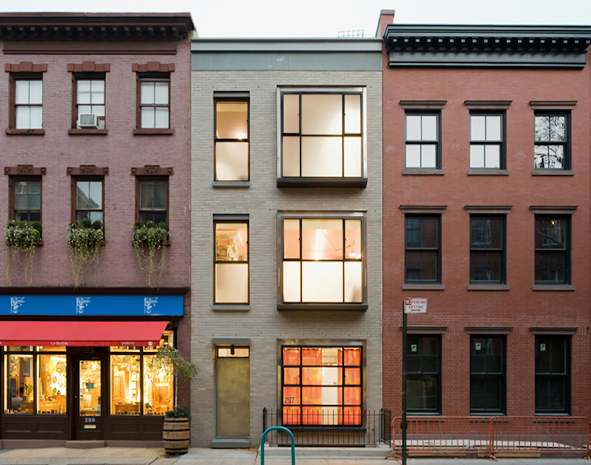 Narrow infill townhouse facade with gray-green exterior, large windows, and warm interior lighting, flanked by traditional brick buildings in New York by Cross Architecture.