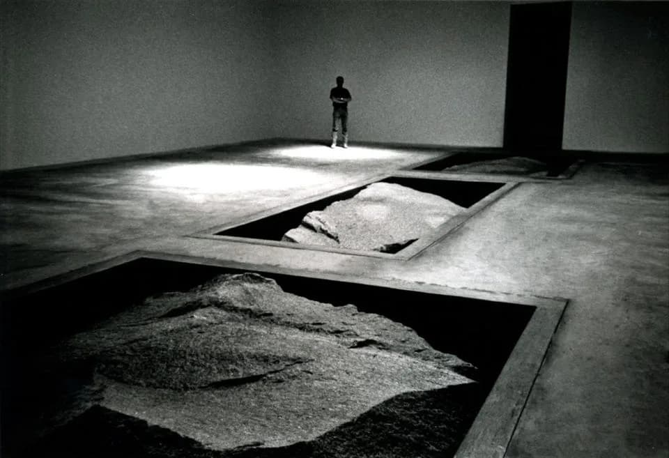 Minimalist concrete installation with geometric cutouts and scattered earth material, featuring dramatic natural lighting and a solitary figure, by Cross Architecture.