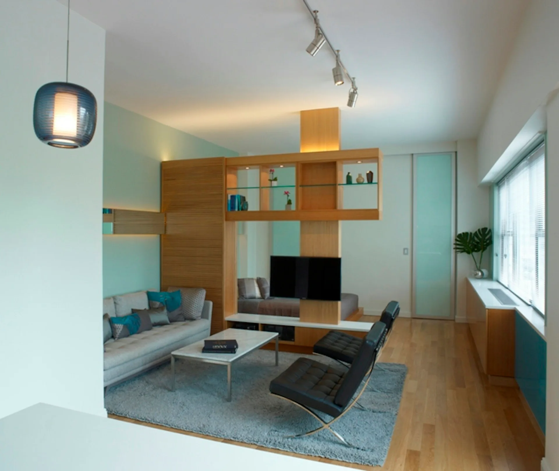 Modern living room with wood and turquoise storage wall, gray sofa, black lounge chair, and wooden flooring, designed by Daedalus Workshop LLC.