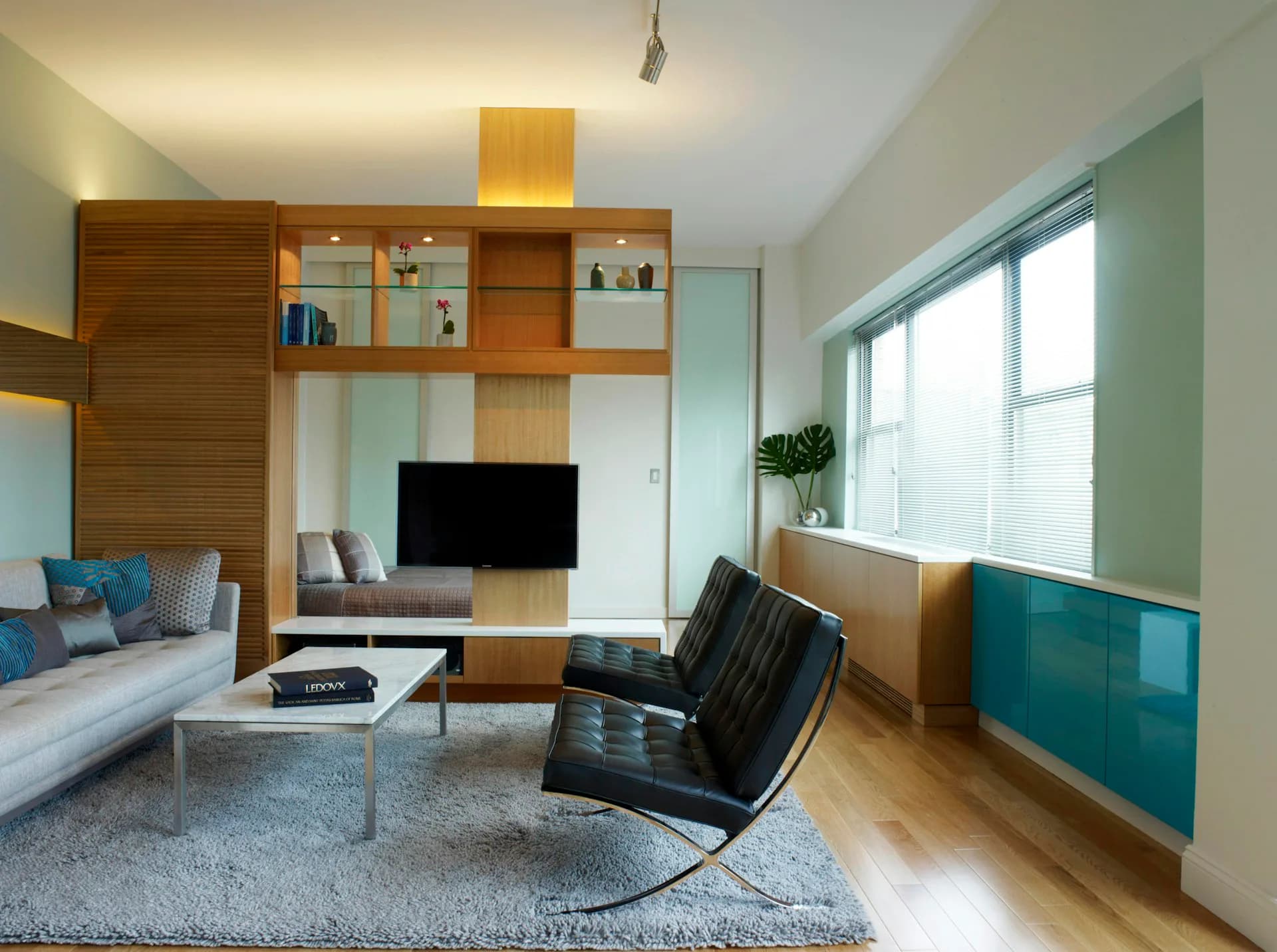 Modern living room with wooden shelving unit, teal cabinetry, and Barcelona chair features synthetic lighting and flawless material presentation by Daedalus Workshop LLC.