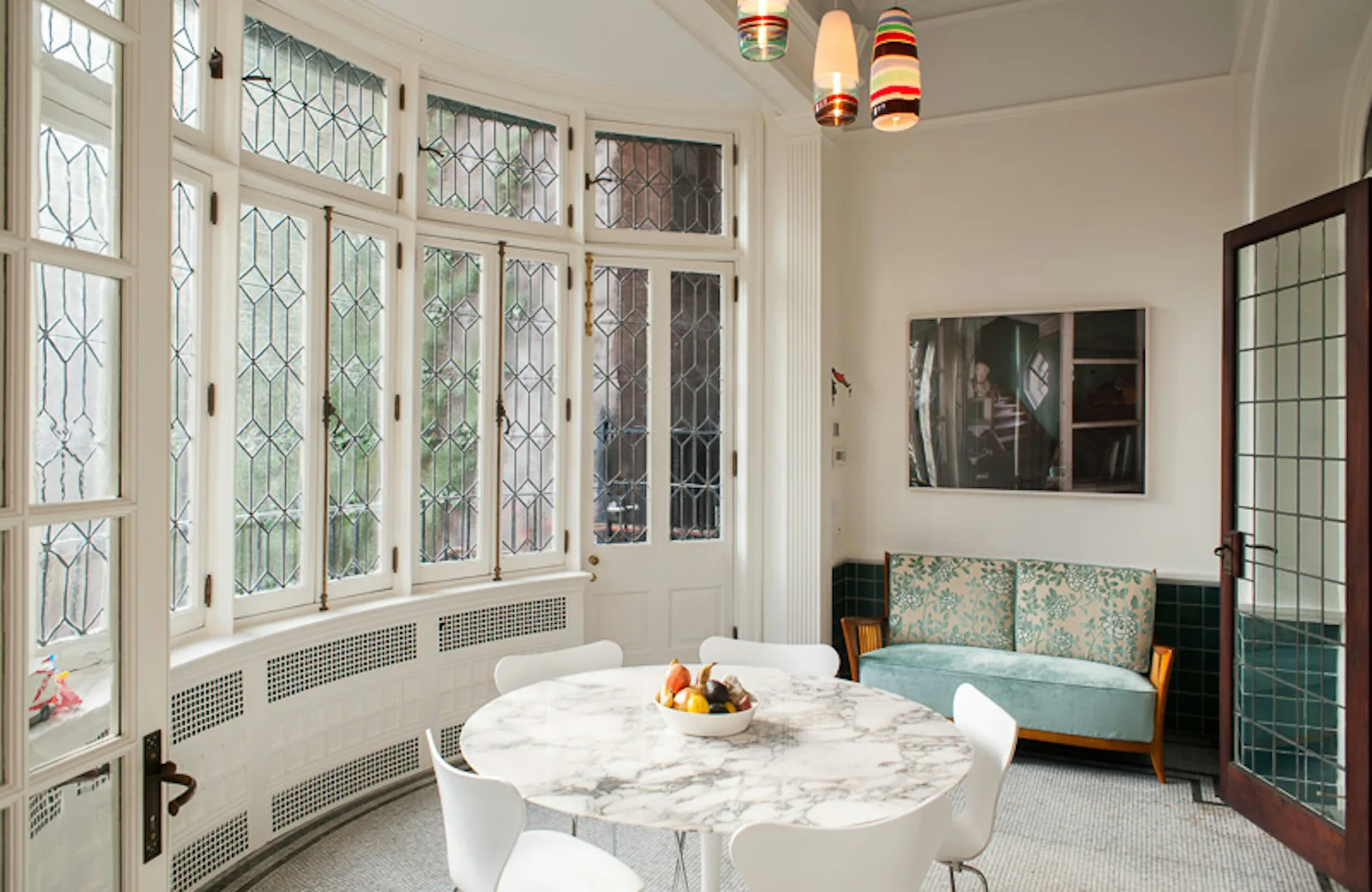 Sunlit sunroom with marble-topped round table, teal upholstered seating, leaded-glass windows, and brass pendant lights by Daniel Frisch Architecture.