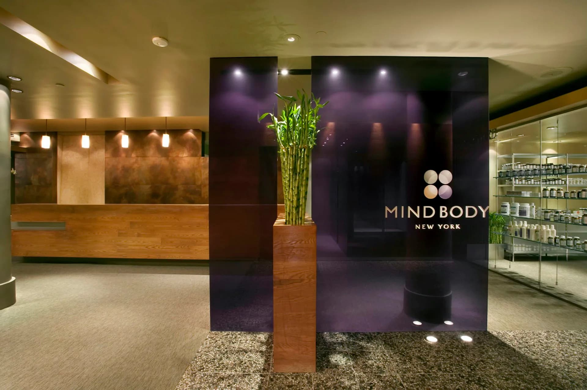 Modern spa reception area with warm wood desk, purple accent wall, bamboo installation, and ambient lighting designed by Daniel Goldner Architects.