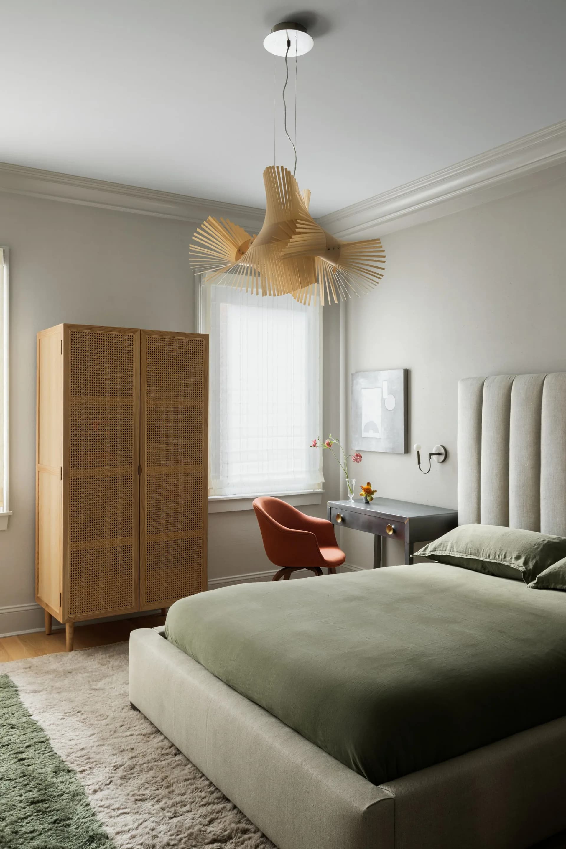 Modern bedroom with sage green bedding, rattan wardrobe, pendant bird sculpture, and upholstered headboard by Danielle Colding Design.