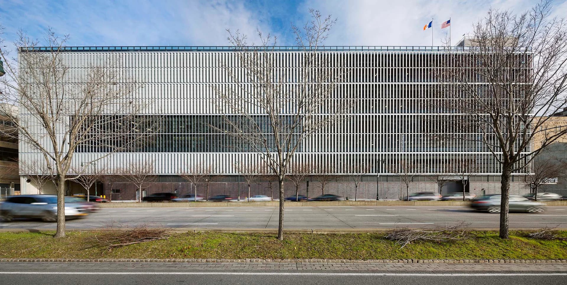 Modern institutional building with horizontal metal louver facade, ground-level parking, bare trees, and manicured lawn, designed by Dattner Architects.
