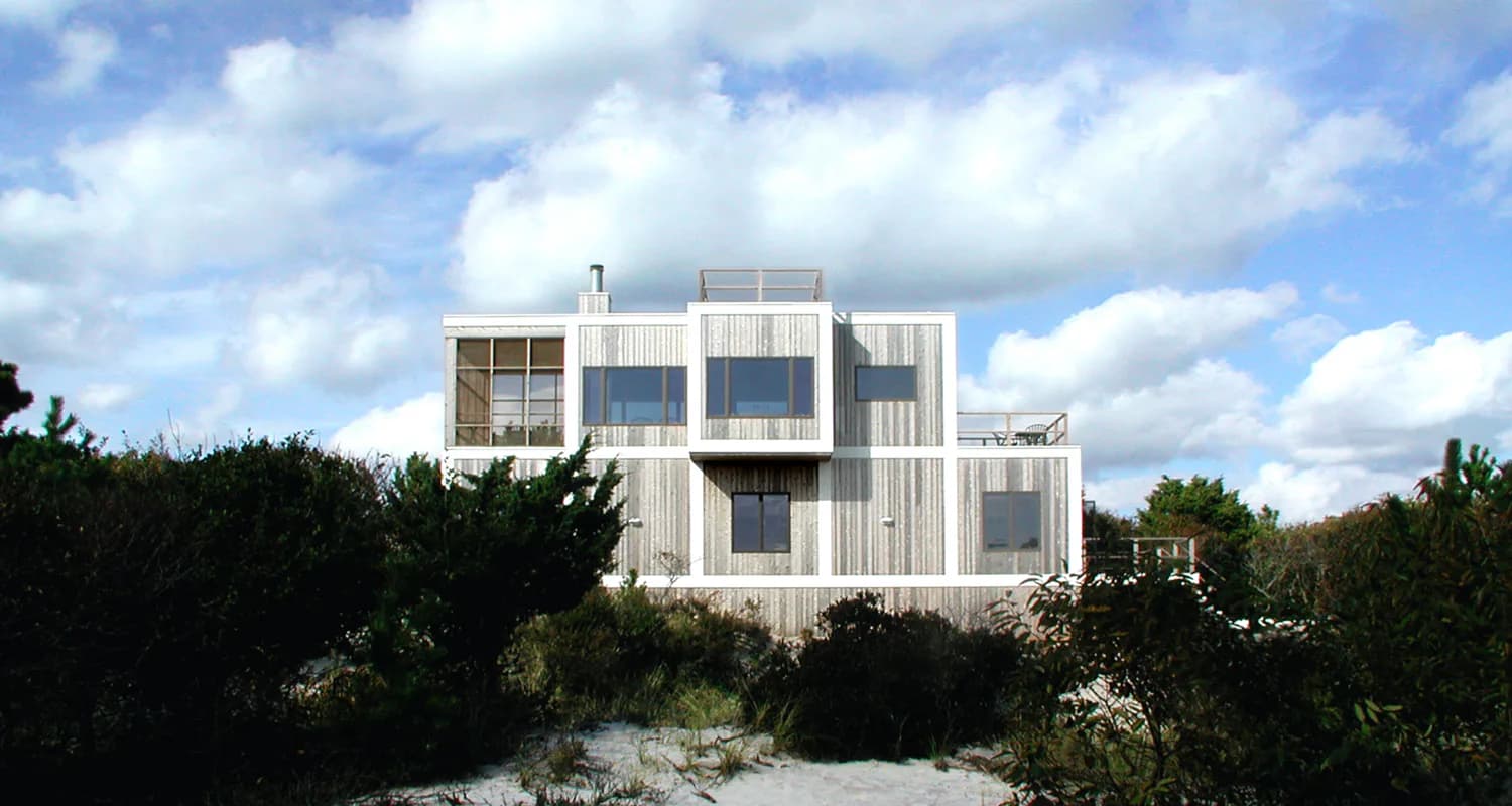Modern beachfront residence with white corrugated metal siding and large window grid, elevated on dunes with vegetation, by David Gauld Architect.