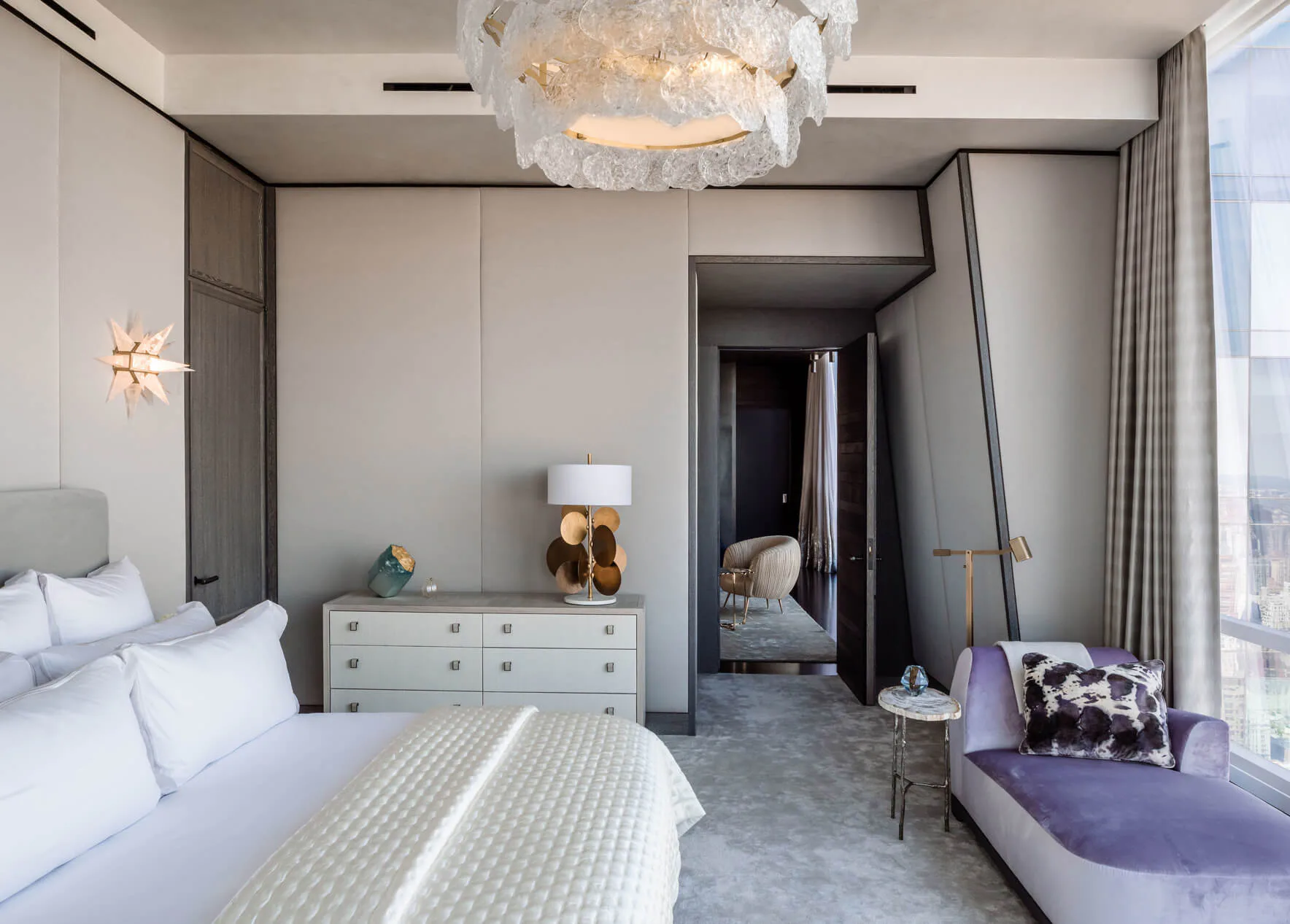 Modern bedroom with cream walls, white bedding, gray-toned furnishings, contemporary chandelier, and floor-to-ceiling windows overlooking a city skyline, designed by DHD Architecture & Interior Design.