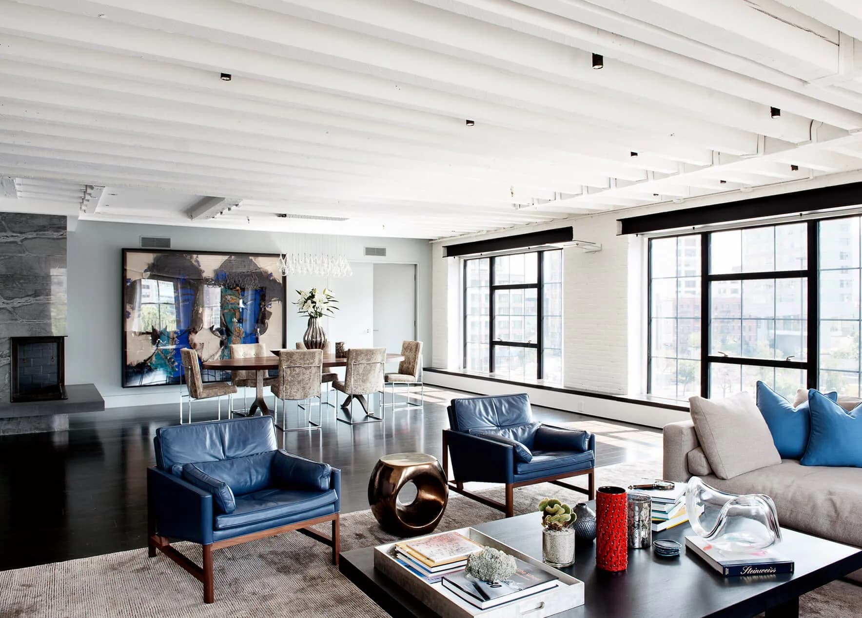 Modern living room with blue velvet chairs, gray sofa, black coffee table, and large windows overlooking city skyline by DHD Architecture & Interior Design.