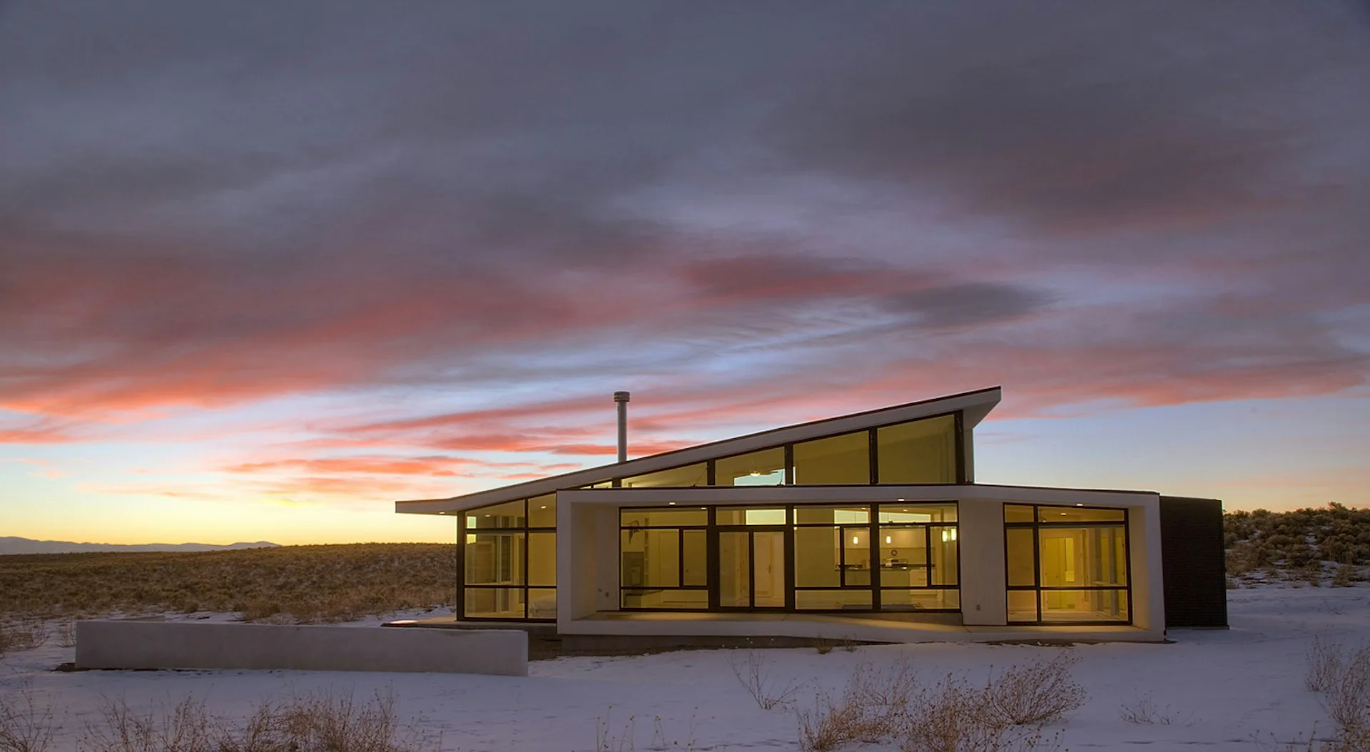 Modern residential structure with angled roof and extensive glazing set in snowy desert landscape at dusk, illuminated from within, by David Jay Weiner, Architects.
