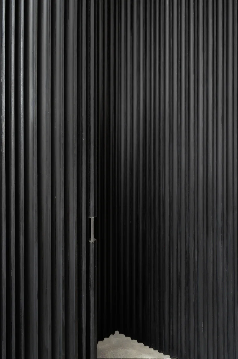 Minimalist interior featuring dramatic vertical corrugated metal wall with strong linear shadows and geometric concrete element below by Davies Toews.
