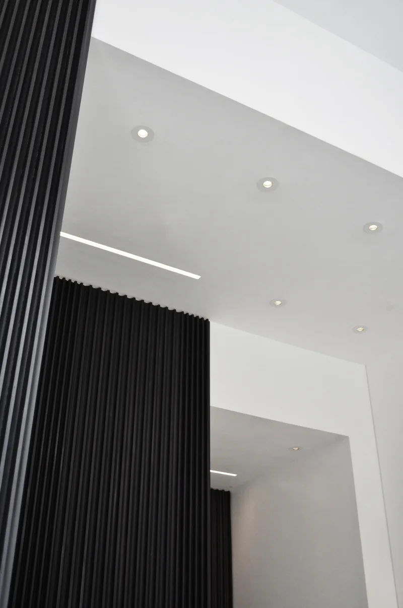 Modern ceiling detail featuring recessed lighting, linear light fixture, and black vertical ribbed wall paneling against white surfaces by Davies Toews.