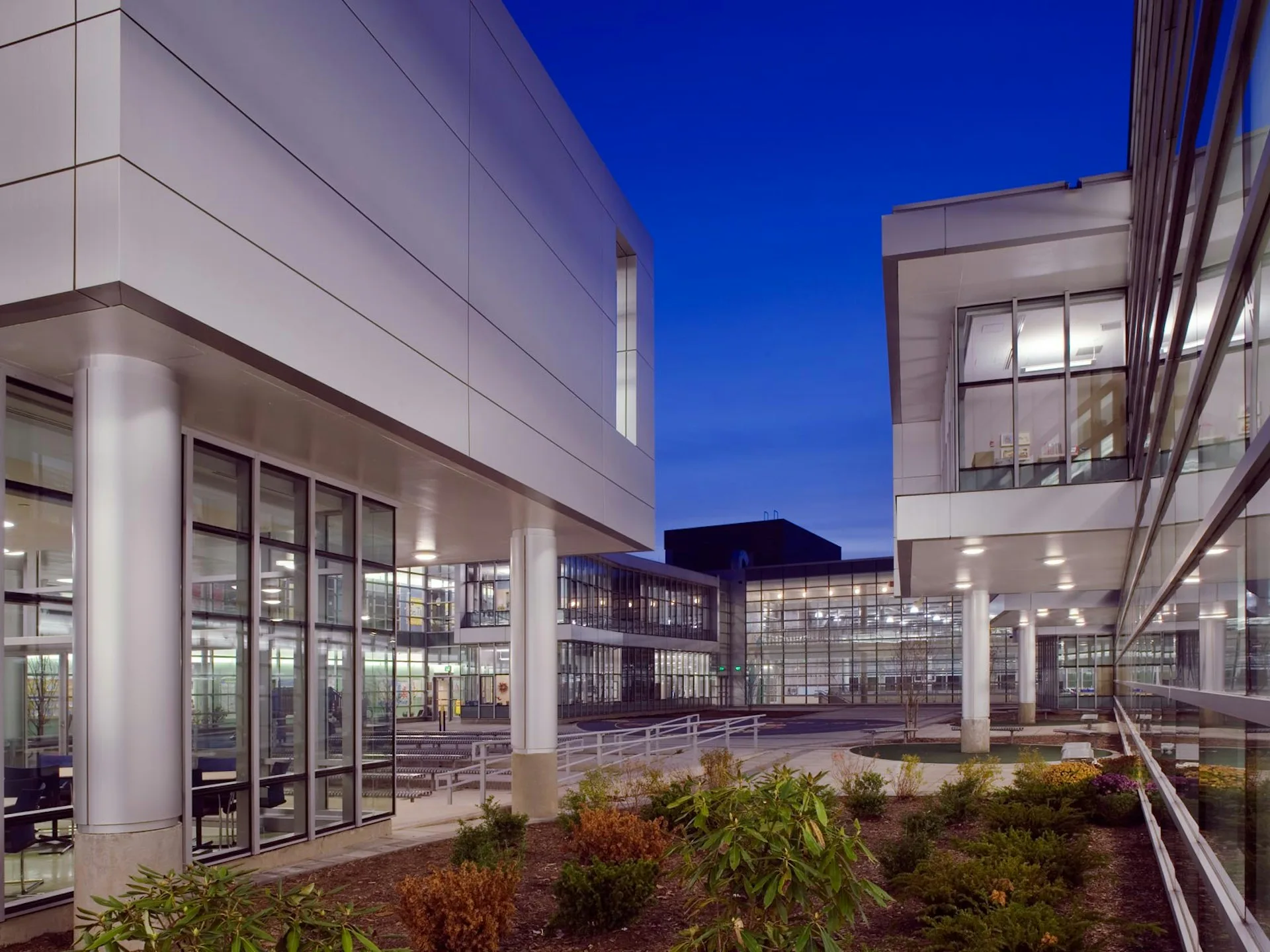 Modern institutional courtyard with illuminated glass-fronted buildings, manicured landscaping, and twilight sky by Davis Brody Bond, a Page Company.