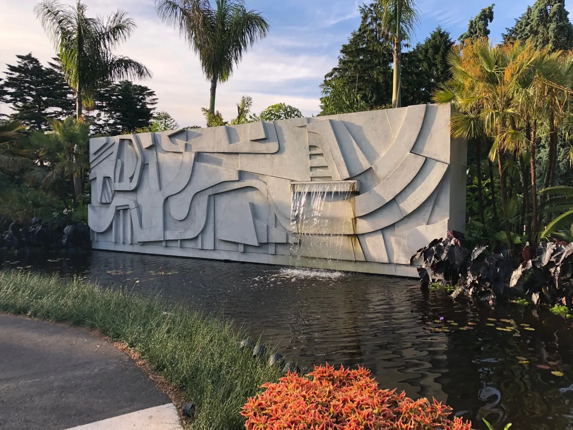 Large-scale white sculptural concrete installation beside a waterway, featuring geometric curved relief patterns, surrounded by tropical palms and landscaping by DCM Fabrication Inc.