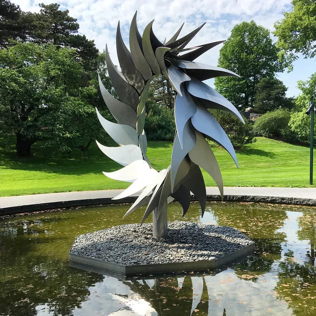 Contemporary metal sculpture installation featuring dynamic curved blades in a reflecting pond, surrounded by manicured lawn and trees, by DCM Fabrication Inc.