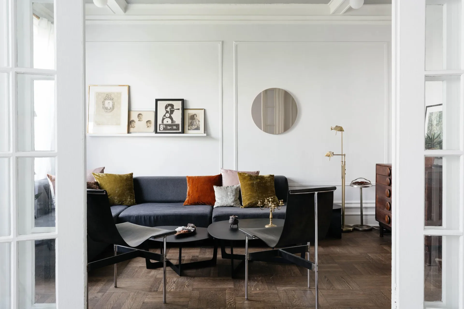 Modern living room with dark navy sofa, mustard and olive accent pillows, glass coffee table, round mirror, and framed artwork on white walls by Delia Kenza Interiors.