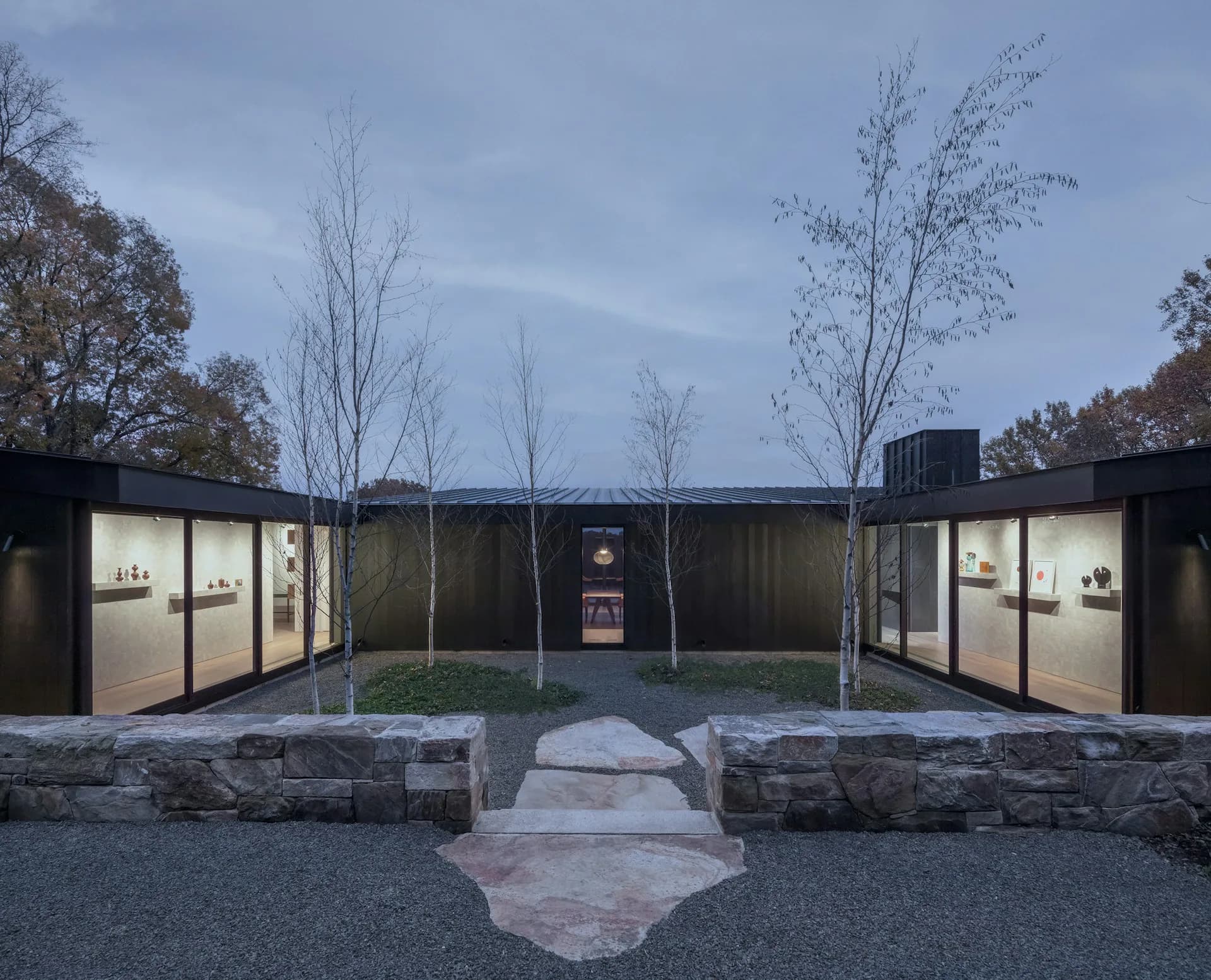 Minimalist pavilion with symmetrical black metal and glass structures flanking a central courtyard, featuring native plantings and stone pathways by Desai Chia Architecture PC.