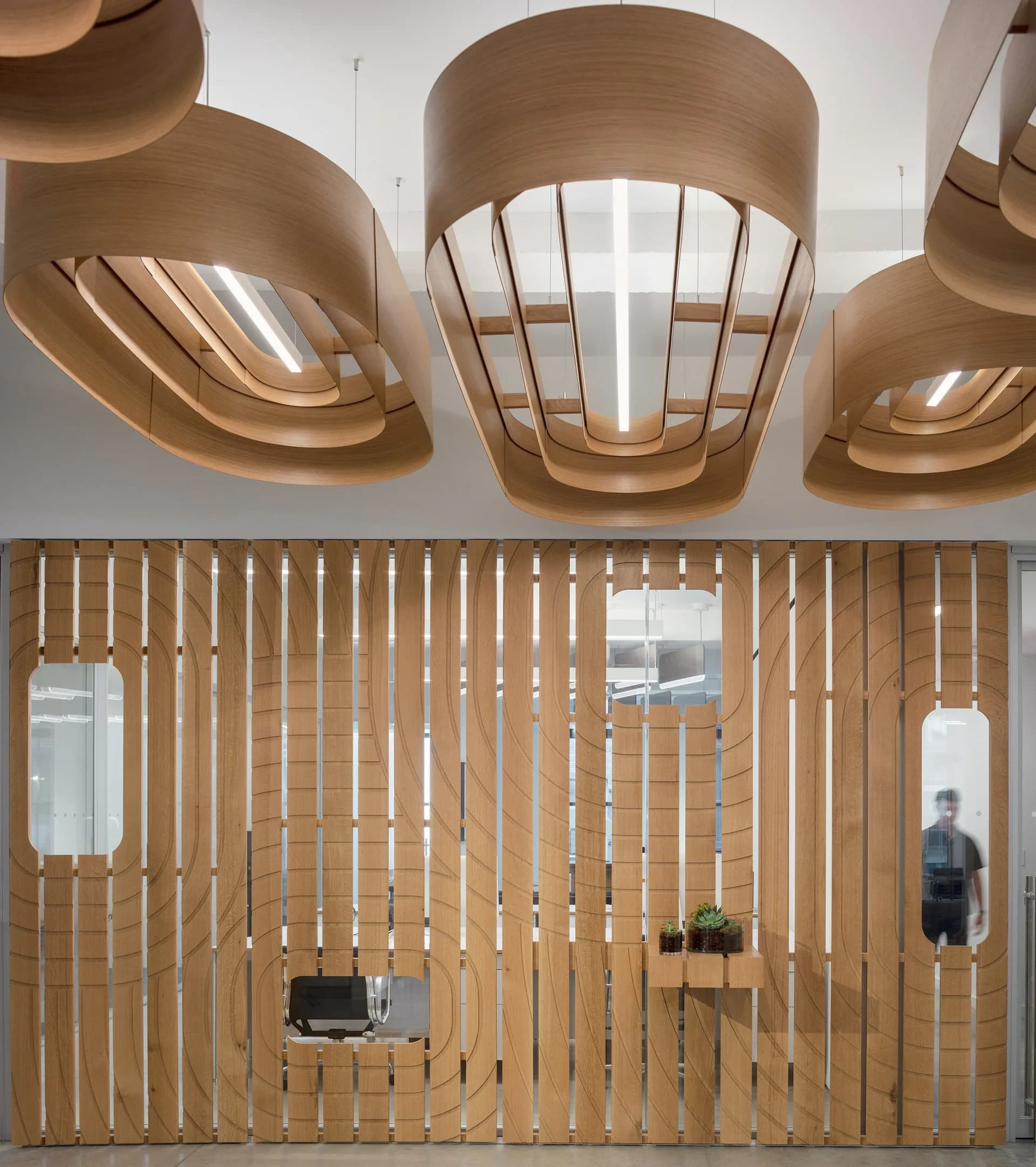 Modern interior with curved wooden pendant lights overhead and vertical wood-slat partition wall with integrated windows, designed by Desai Chia Architecture PC.