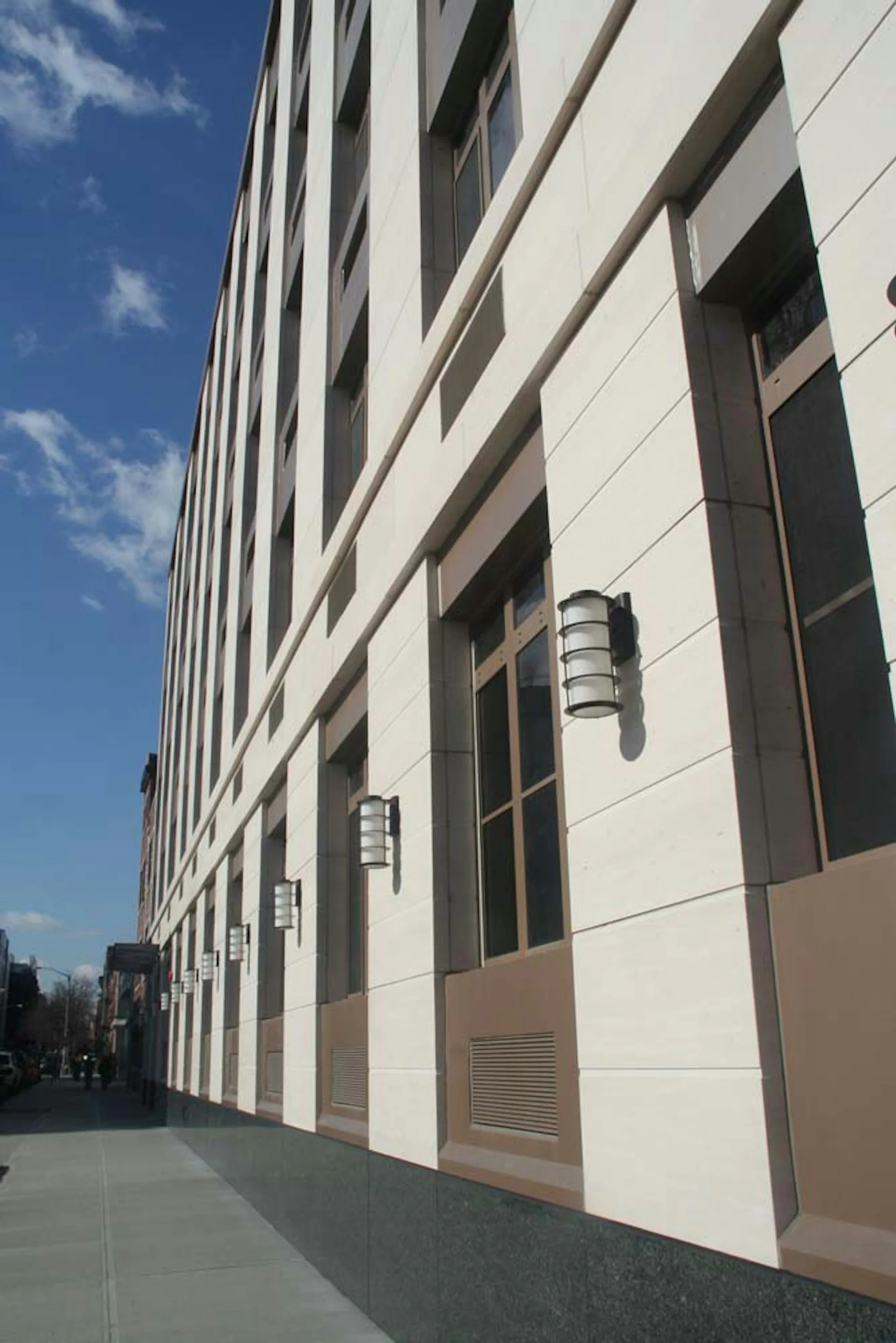 Modern mid-rise building facade featuring white and tan panels with dark vertical elements and cylindrical wall sconces, photographed in daylight by Design AIDD Architecture, DPC.