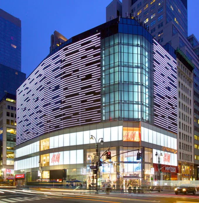 Curved retail building facade with dramatic horizontal LED lighting bands illuminating white and dark striations, glowing storefront windows, and glass cylindrical tower element by Design One Lighting.