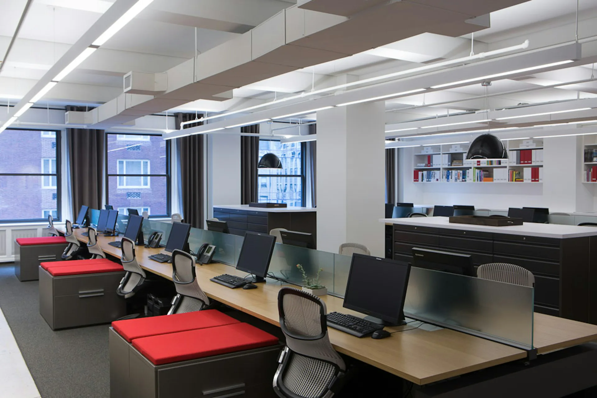 Modern open-plan office with natural light, featuring red accent desks, black ergonomic chairs, natural wood surfaces, and glass partitions, designed by Design Republic.