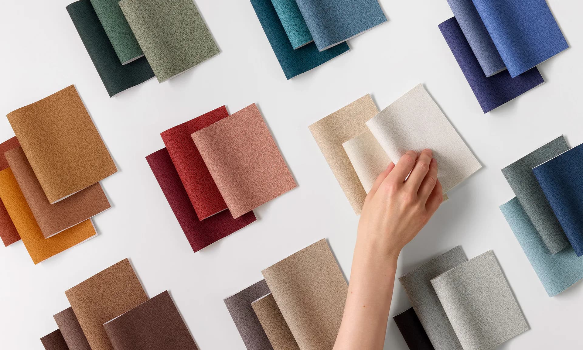 Organized color swatches in warm, cool, and neutral tones arranged on white background, with a hand selecting a cream sample, showcasing Designtex's fabric palette.