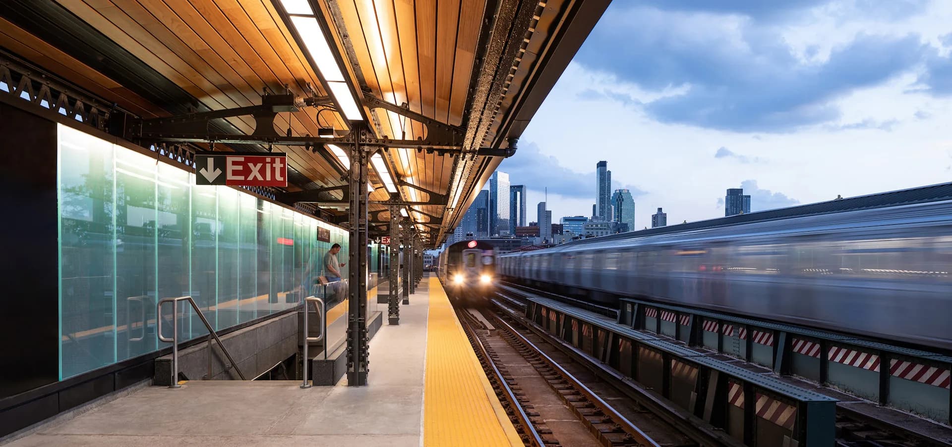 Modern subway station platform with wooden ceiling, glass enclosure, yellow safety edge, and arriving train, designed by di Domenico + Partners, LLP.