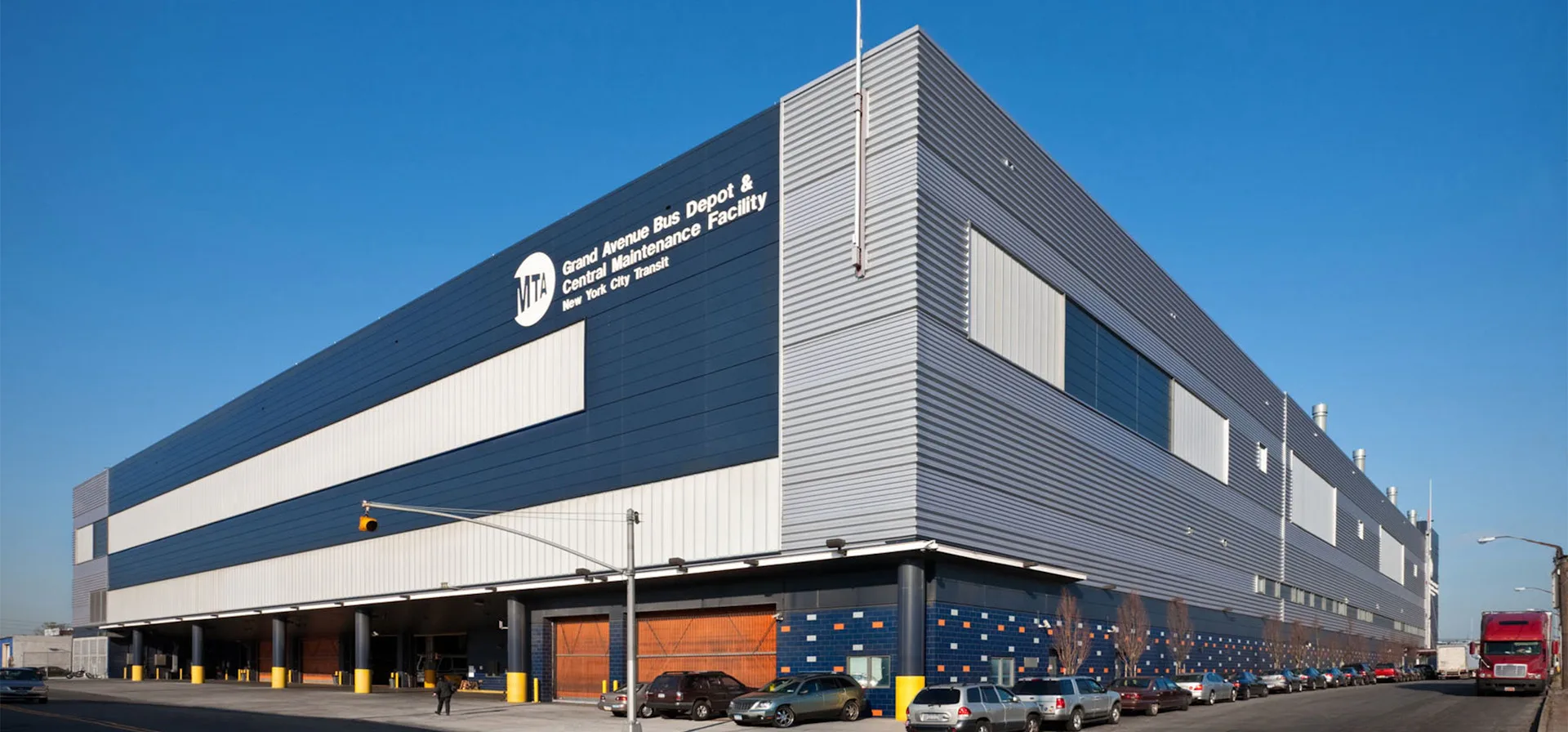 Modern industrial warehouse building with dark blue and gray corrugated metal facade, horizontal striping, and ground-level orange garage doors designed by di Domenico + Partners, LLP.