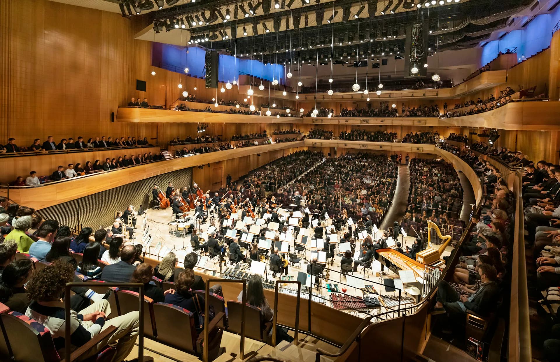 A concert hall with warm wood interiors, tiered seating galleries, suspended lighting, and a full orchestra on stage, designed by Diamond Schmitt.