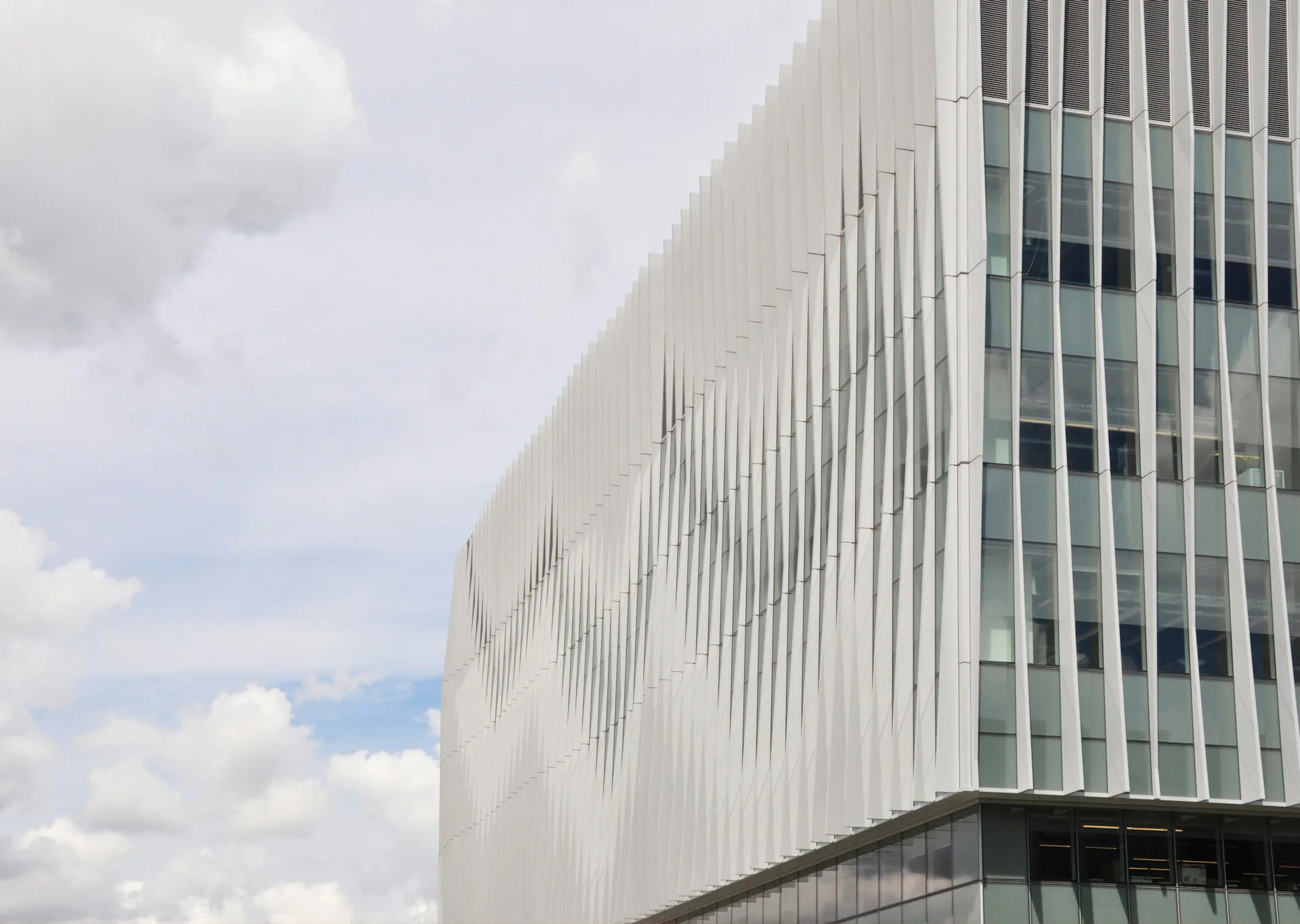 Modern institutional building facade with vertical ribbed white metal cladding and glass panels against a cloudy sky, by Diamond Schmitt.