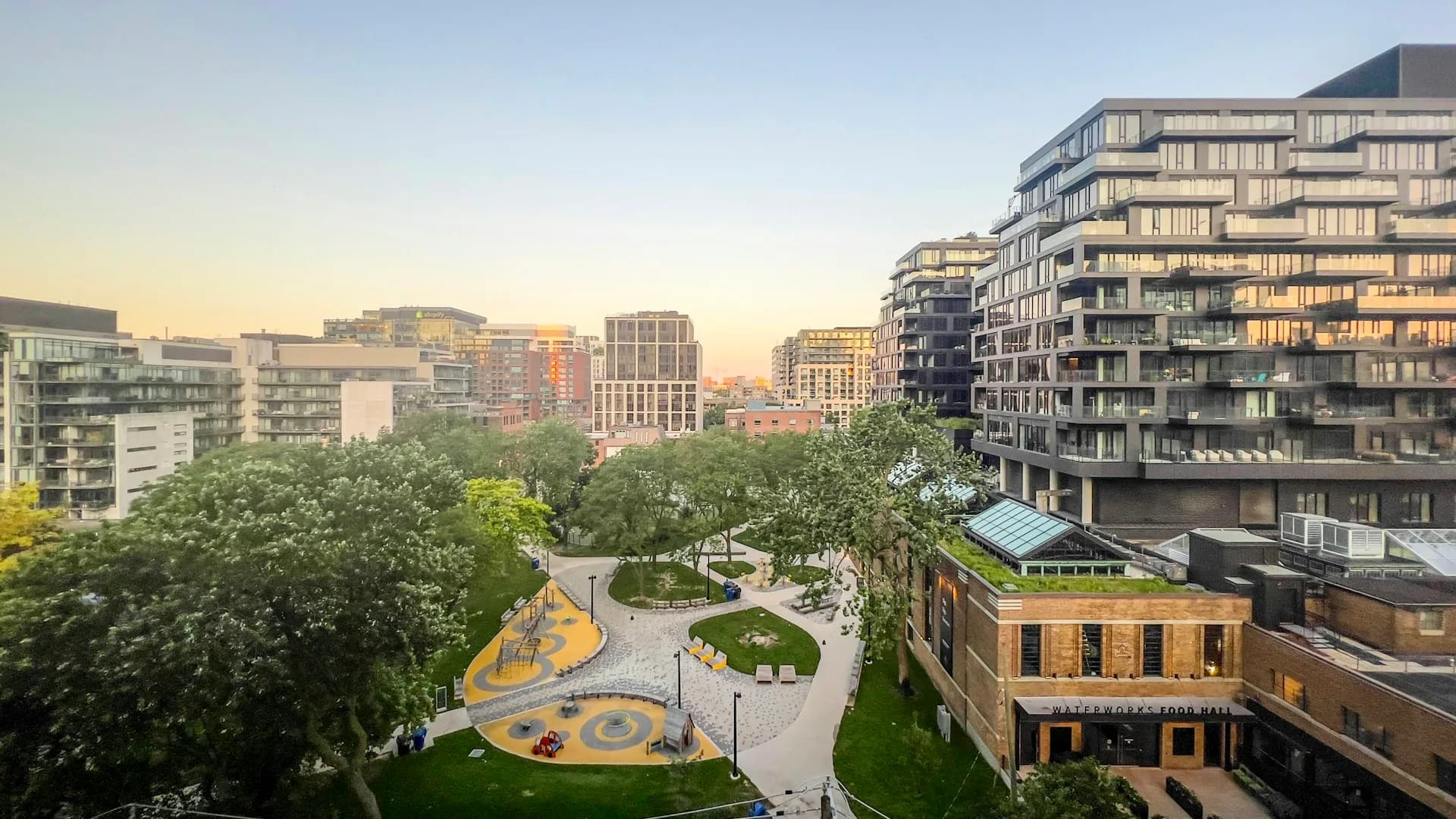 Urban plaza with curved yellow sculptural elements and mature trees, surrounded by contemporary residential and mixed-use buildings at dusk by Diamond Schmitt.