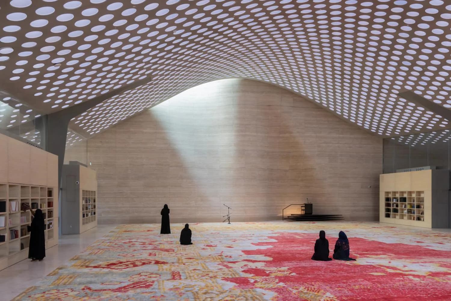 Vast concrete prayer hall with geometric perforated ceiling, colorful patterned carpet, and silhouetted figures by Diller Scofidio + Renfro.