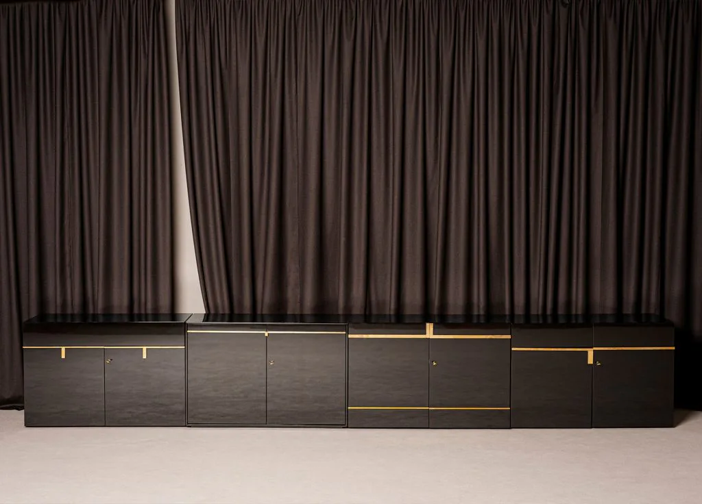 Minimalist dark cabinetry with brass accents and integrated lighting beneath heavy draped curtains, by Dimoremilano.