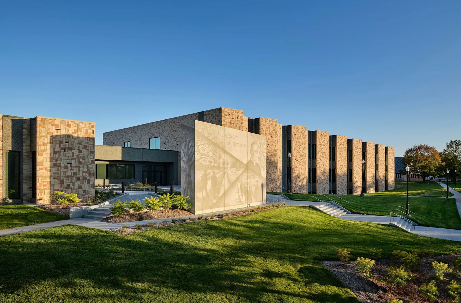 Modern institutional building with geometric black and tan striped facade, featuring expansive glass frontage and manicured landscaping with green lawn by DLR Group.