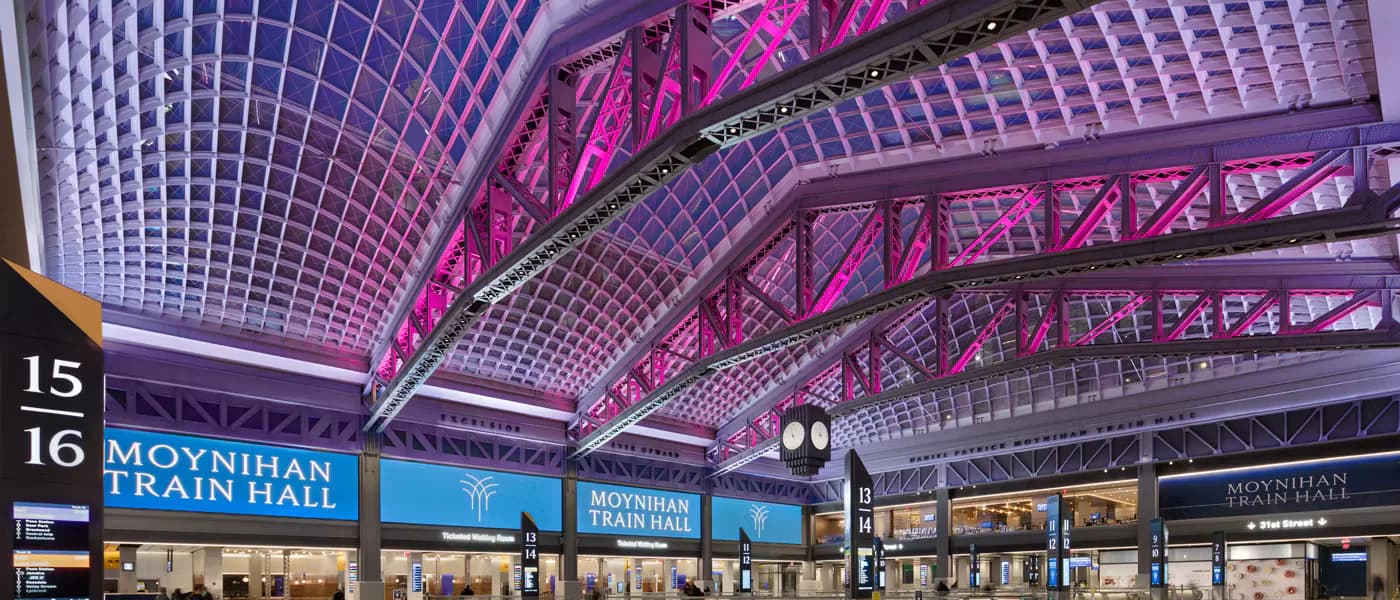 Moynihan Train Hall interior featuring vibrant pink structural beams, geometric ceiling grid, and modern signage in a renovated transit station by Domingo González Associates (DGA).