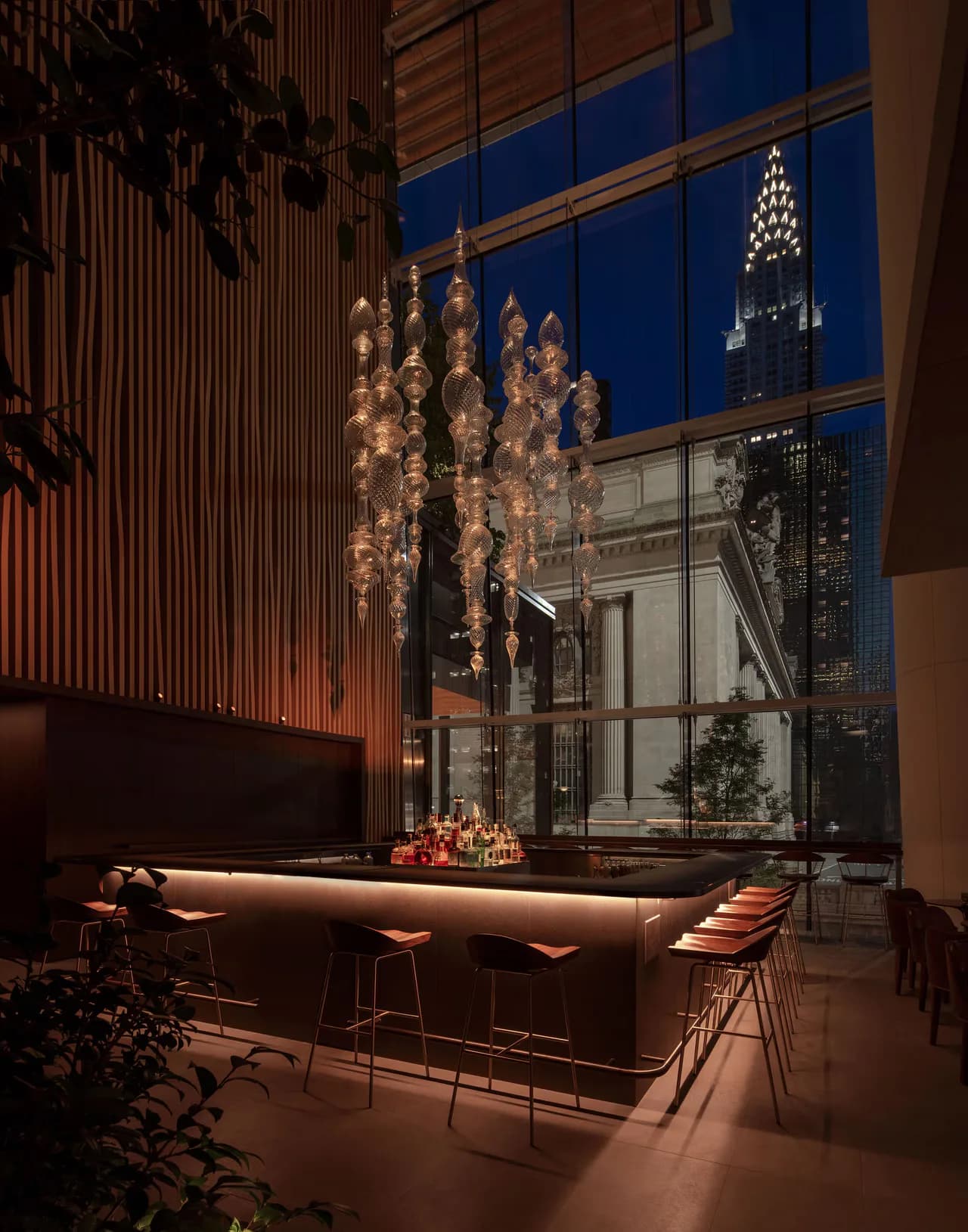 Modern bar interior with warm wood paneling, illuminated pendant lights, and floor-to-ceiling windows framing NYC skyline at dusk by Dot Dash.