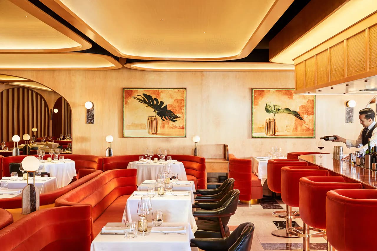 Luxurious restaurant dining room with curved red velvet banquettes, white tablecloths, botanical artwork, and warm ambient lighting designed by Dot Dash.