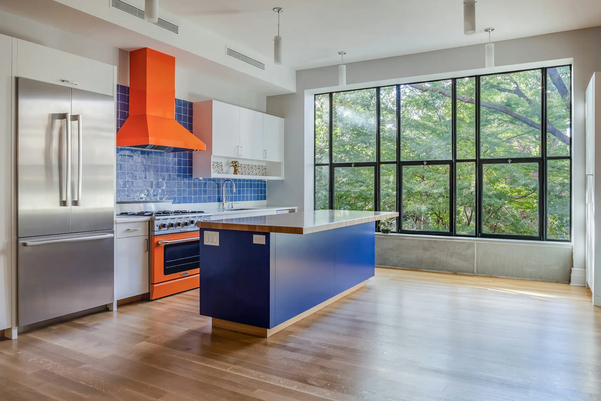 Modern kitchen with orange hood, blue tile backsplash, blue island, and large multi-pane windows overlooking greenery by Ed Kopel, Architect, P.C.