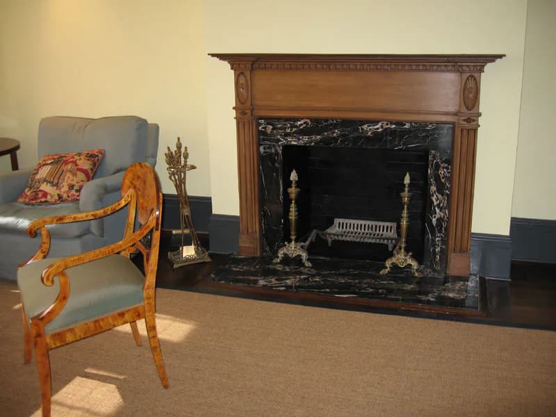 Elegant living room interior featuring a wooden fireplace mantel with brass andirons, a blue upholstered wingback chair with orange carved details, and warm cream and dark blue wall palette by Edward Dew Architect.
