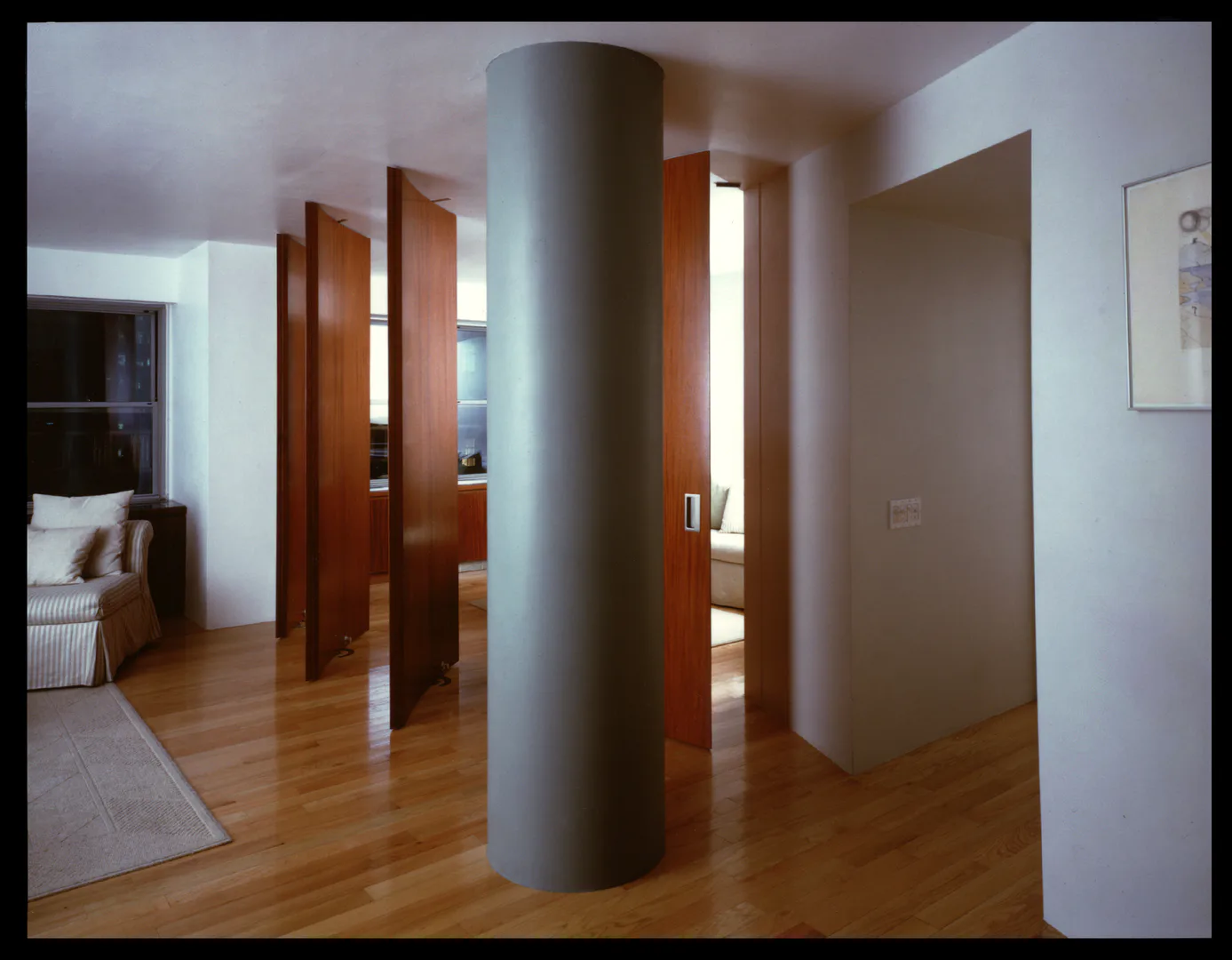 Modern hallway with wooden flooring, white walls, and a distinctive cylindrical gray column, featuring dark wood doors opening to adjacent spaces, designed by Edward Dew Architect.