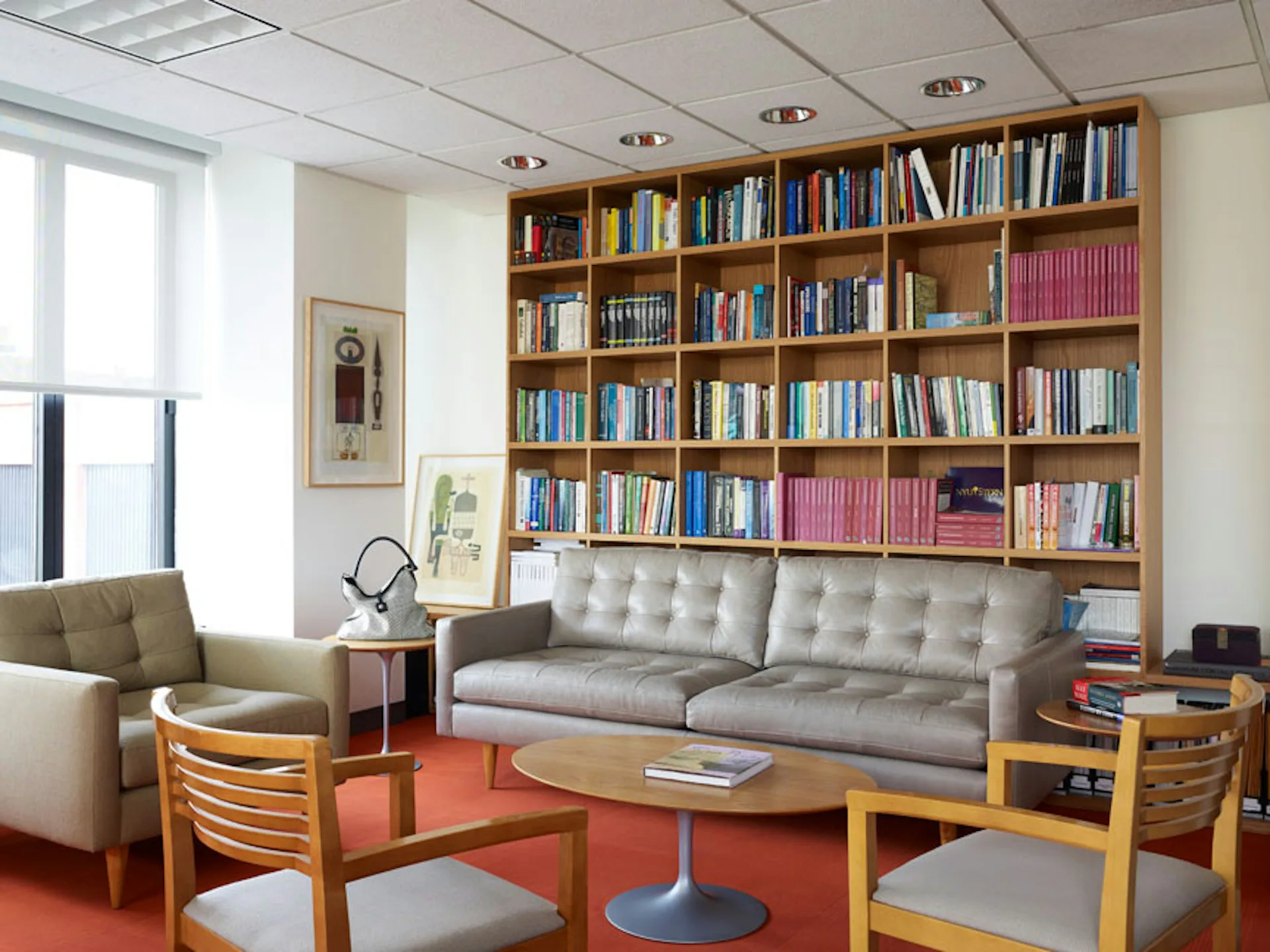 Modern library lounge with built-in wooden bookshelves, gray upholstered sofa, mid-century wooden chairs, and orange rug by EFM Design.