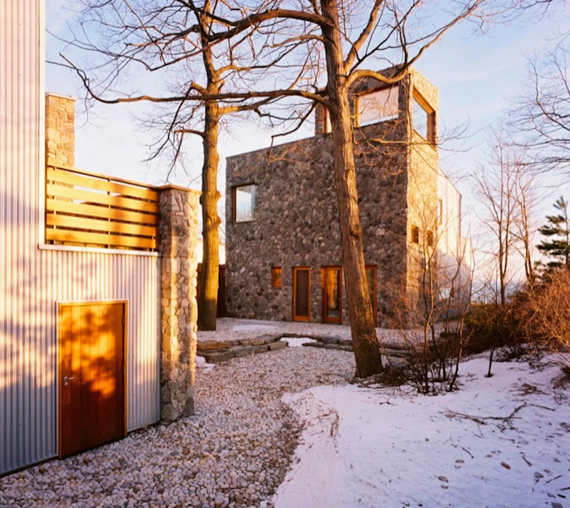 Contemporary stone and metal structure nestled among bare trees in a winter landscape, featuring geometric forms and warm golden hour lighting by EFM Design.